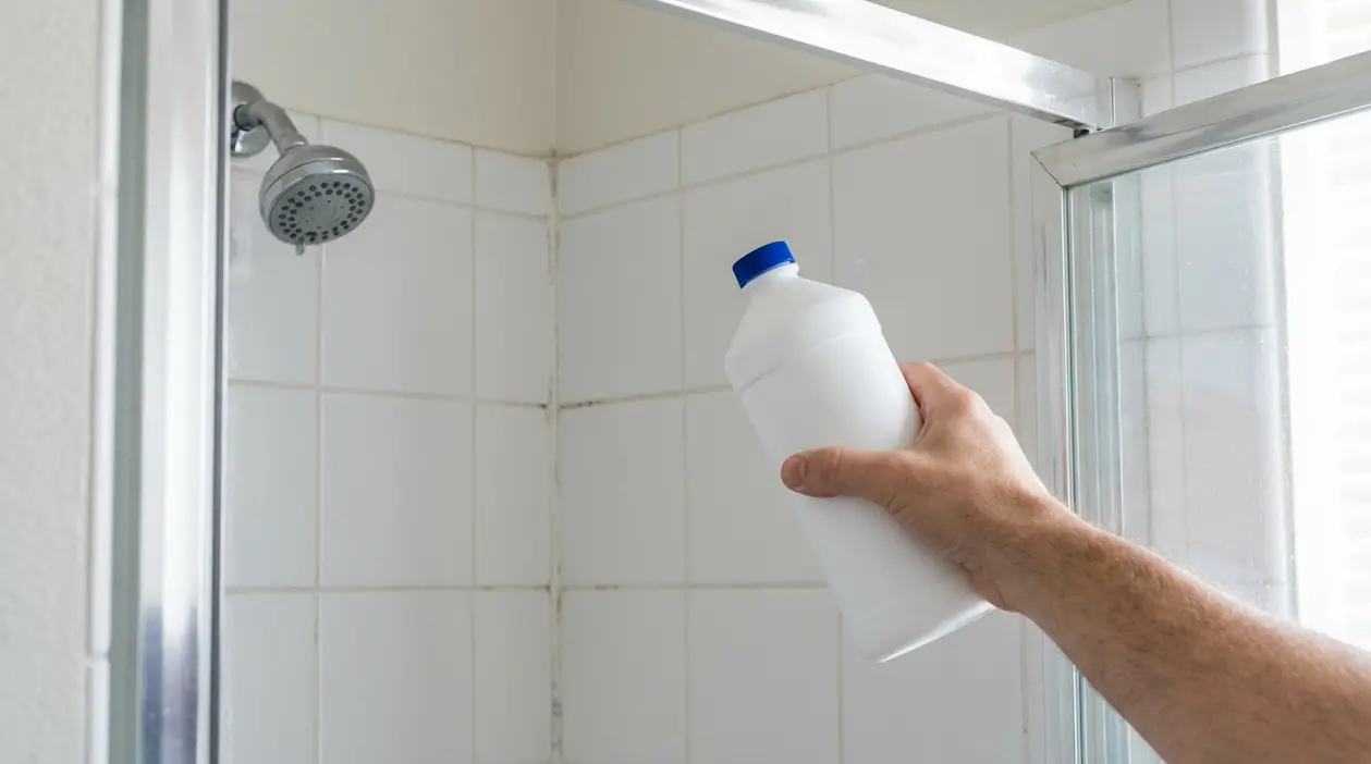 Mano che tiene un flacone di acqua ossigenata nella doccia davanti a piastrelle bianche e soffione