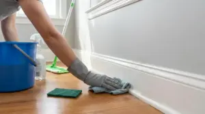 Persona con guanto che pulisce i battiscopa bianchi con un panno in una stanza luminosa