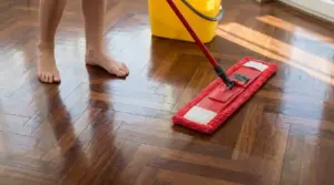 Persona che passa un panno mop su un parquet lucido in casa, accanto a un secchio giallo