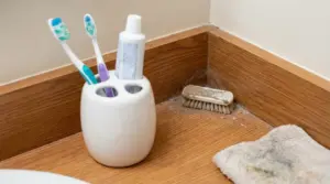 Angolo del bagno con polvere sul battiscopa, spazzolina, panno e portaspazzolini sul piano in legno