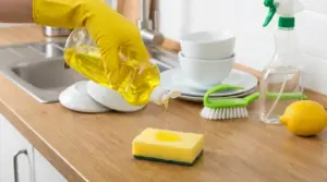 Mano con guanto versa detersivo per piatti su una spugna sul piano cucina vicino al lavello e a piatti impilati