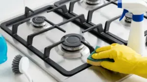 Mano con guanto che pulisce i bruciatori del piano cottura a gas in acciaio con spugna e detergente spray