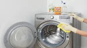 Persona con guanti pulisce una lavatrice a carica frontale aperta con panno e detergenti come aceto e bicarbonato
