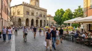 Persone a passeggio e in bici in una piazza storica italiana con caffè all'aperto e palazzo comunale