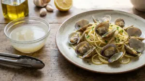 Piatto di spaghetti alle vongole con prezzemolo su tavolo rustico, accanto a una ciotola di brodo e olio d'oliva.