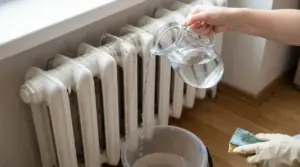 Mano che versa acqua da una brocca su un termosifone sporco, con secchio e spugna per la pulizia