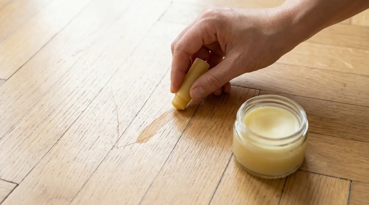 Una mano applica un rimedio naturale su un parquet graffiato con un barattolo di cera accanto