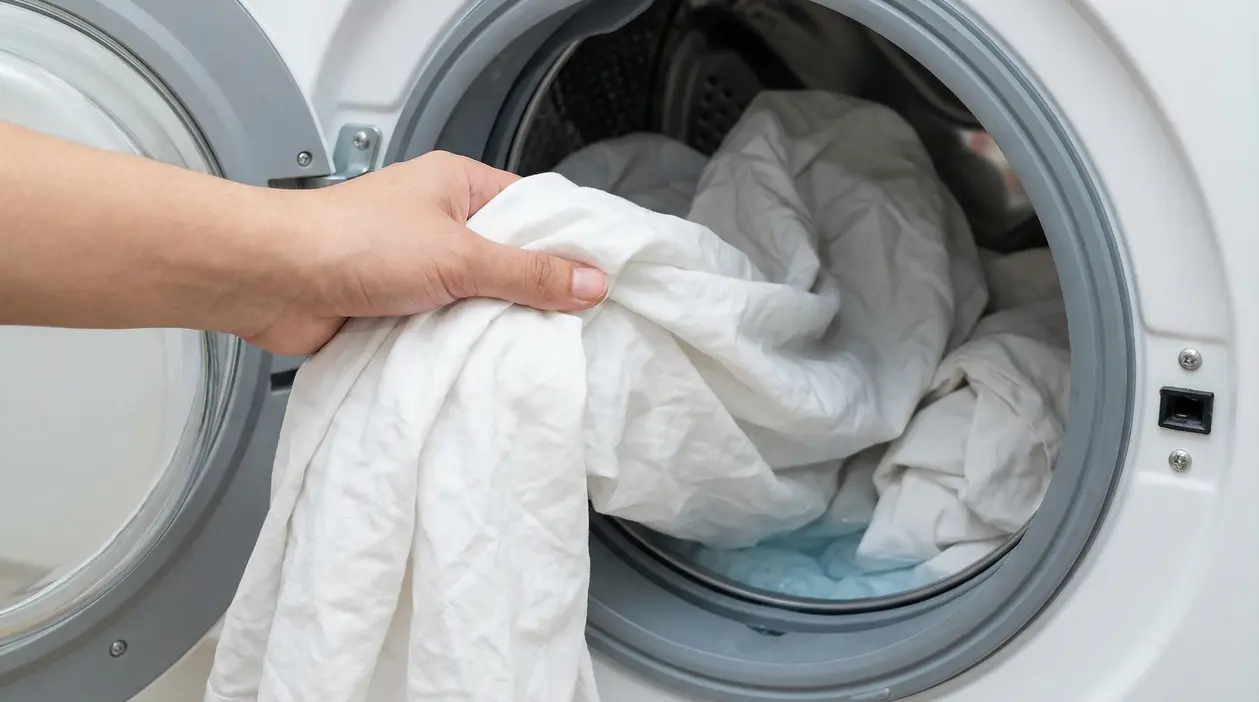 Una mano inserisce lenzuola bianche nella lavatrice a carica frontale per un lavaggio più igienico